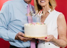 An elderly couple joyfully holding a 'Mr & Mrs' wedding anniversary cake.